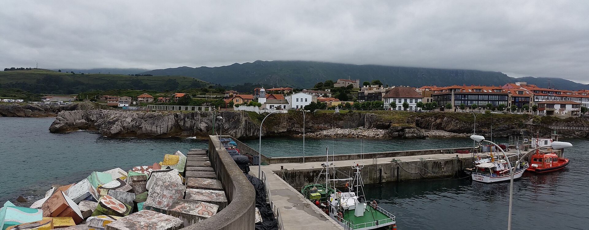 Vista panorámica del puerto de Llanes con los Cubos de la Memoria en el espigón, barcos de pesca amarrados y la villa con la sierra al fondo.